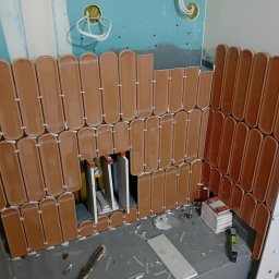 Firma Budowlano-Instalacyjna Bartłomiej Kopacz - Układanie podłużnych, brązowych płytek w łazience. Widoczne krzyżyki dystansowe i fragmenty płytek. Etap prac wykończeniowych na ścianie.