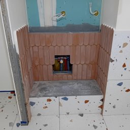 Firma Budowlano-Instalacyjna Bartłomiej Kopacz - Narożnik łazienki w trakcie wykańczania: ułożone cegiełki i terazzo, widoczne rury i instalacja elektryczna. Metalowy profil w rogu.