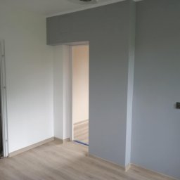 Firma Budowlano-Instalacyjna Bartłomiej Kopacz - Wnętrze po malowaniu: szara ściana z kontaktem, biała ściana, drzwi w tle i panele podłogowe. Widoczny fragment sufitu z oświetleniem.