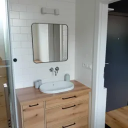 Nowoczesna łazienka z białymi płytkami, drewnianą szafką pod umywalką i czarnym lustrem. Geometryczna podłoga. Widoczny fragment kabiny prysznicowej.