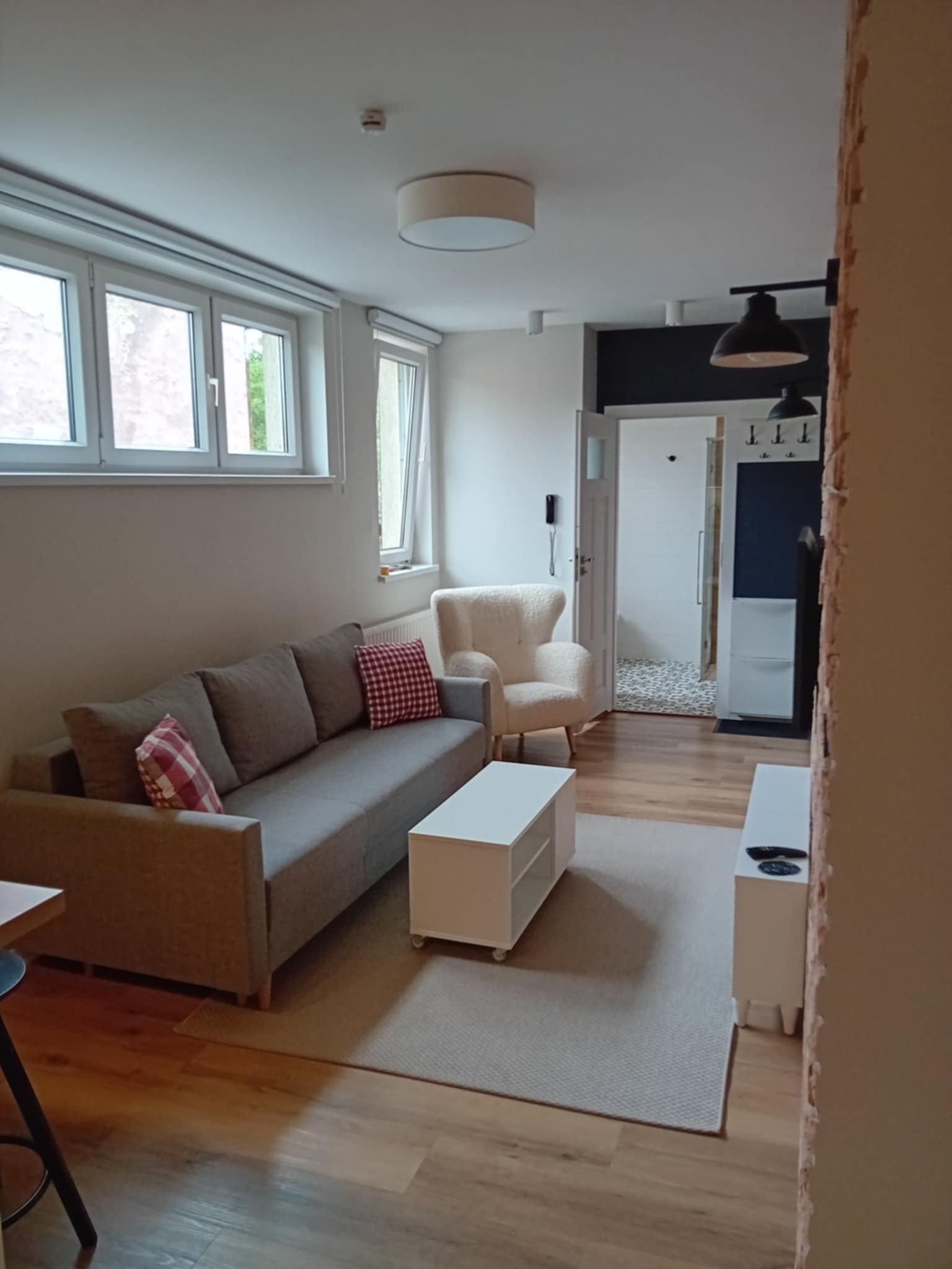 Jasne wnętrze pokoju dziennego z szarą sofą, białym fotelem i stolikiem kawowym. Widoczne okna i drzwi do łazienki z wzorzystą podłogą. Ściana z widocznymi śladami prac remontowych.