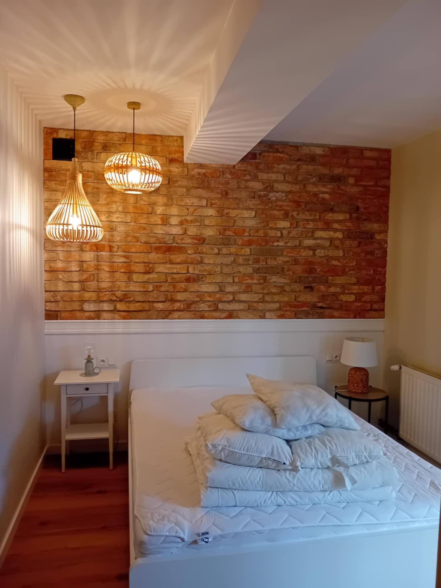 Ustawne wnętrze sypialni z ceglaną ścianą, lampami z wikliny i białymi meblami. Widok na łóżko z pościelą i stolik nocny z lampką.