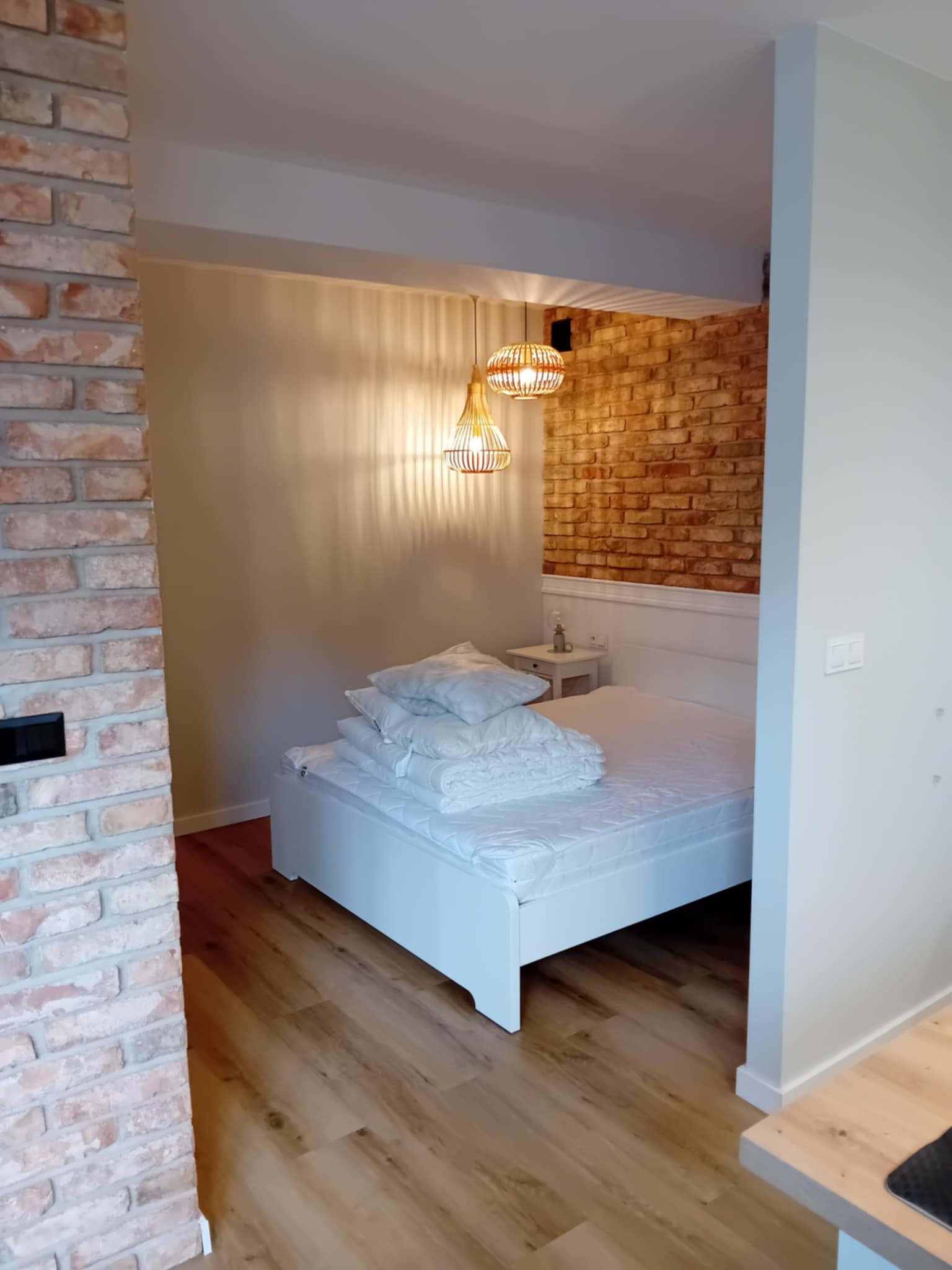 Wykończona sypialnia z ceglaną ścianą, drewnianą podłogą i białym łóżkiem. Dwa wiklinowe abażury zwisają z sufitu, oświetlając wnętrze. Widok zza ceglanego filaru.