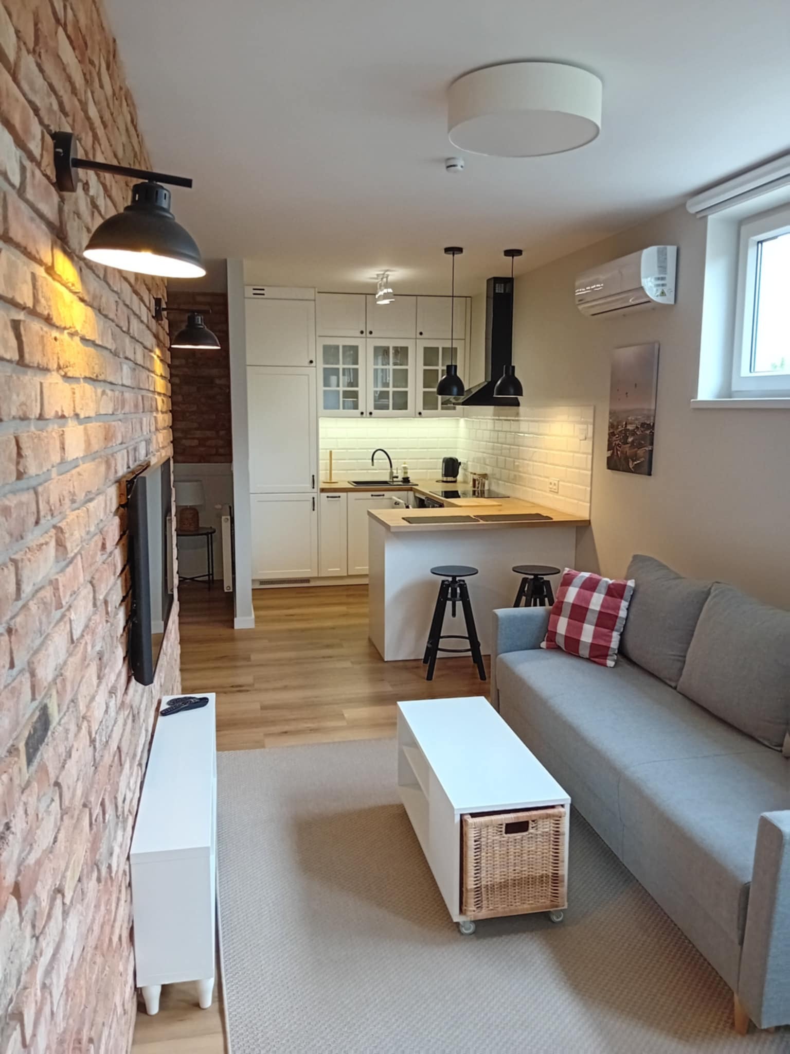Salon z aneksem kuchennym: ceglana ściana z lampami, szara sofa, białe meble kuchenne i wyspa z hokerami. Widoczne detale wykończenia wnętrza.
