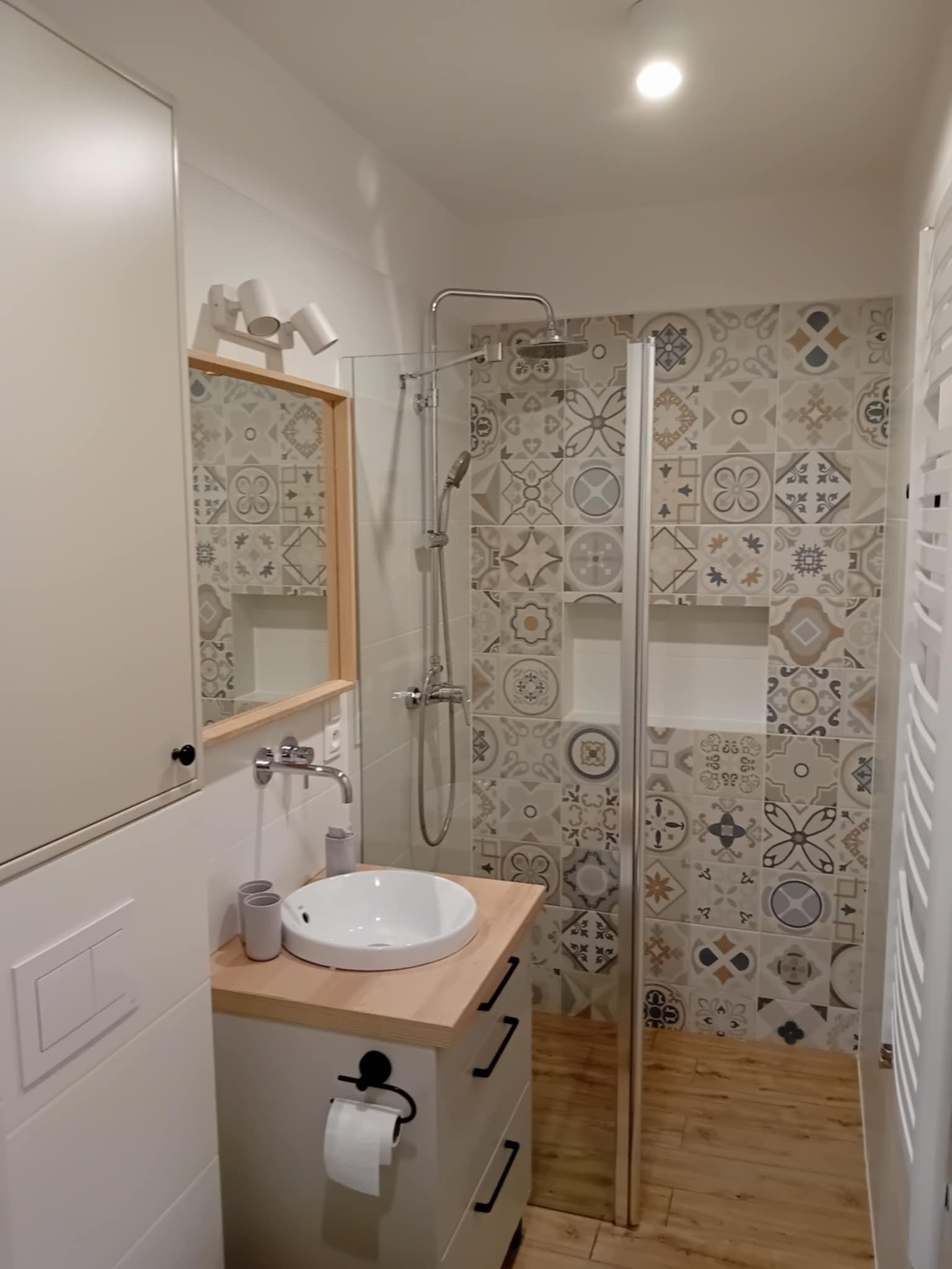 Nowoczesna łazienka z kabiną prysznicową, umywalką nablatową i dekoracyjnymi płytkami w odcieniach szarości i beżu. Jasne drewno blatu i podłogi dodaje ciepła.