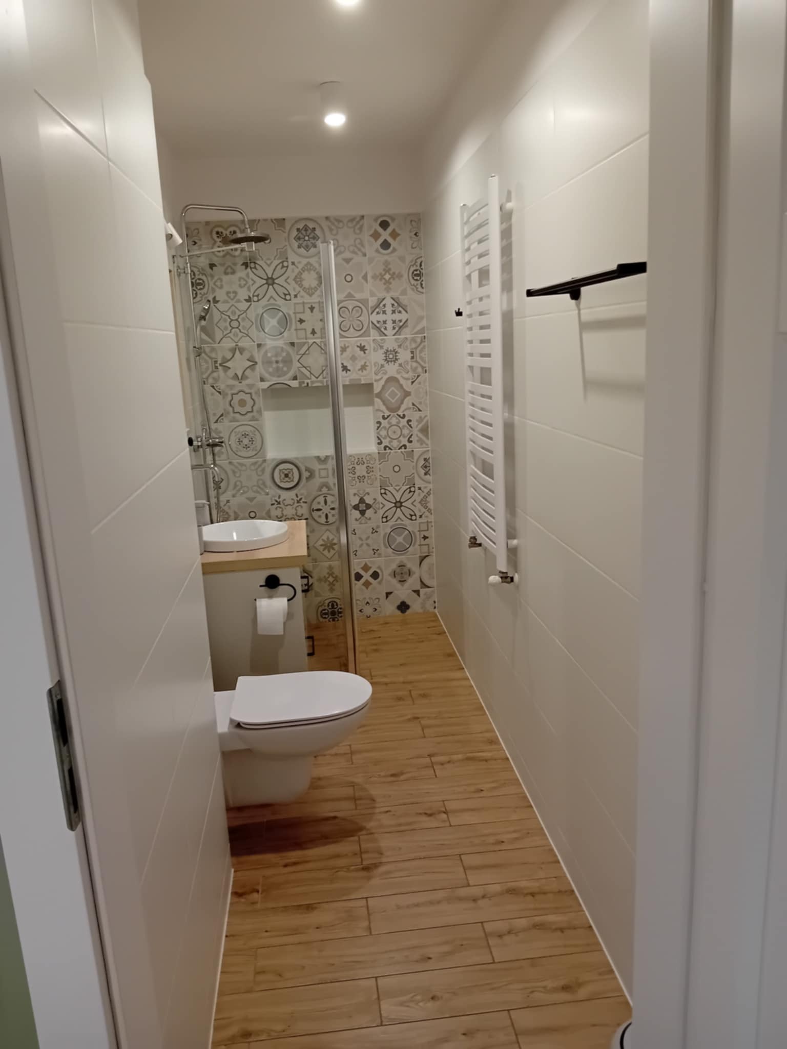 Jasna łazienka z kabiną prysznicową, umywalką nablatową i grzejnikiem drabinkowym. Na ścianach płytki w geometryczne wzory i imitujące drewno na podłodze.