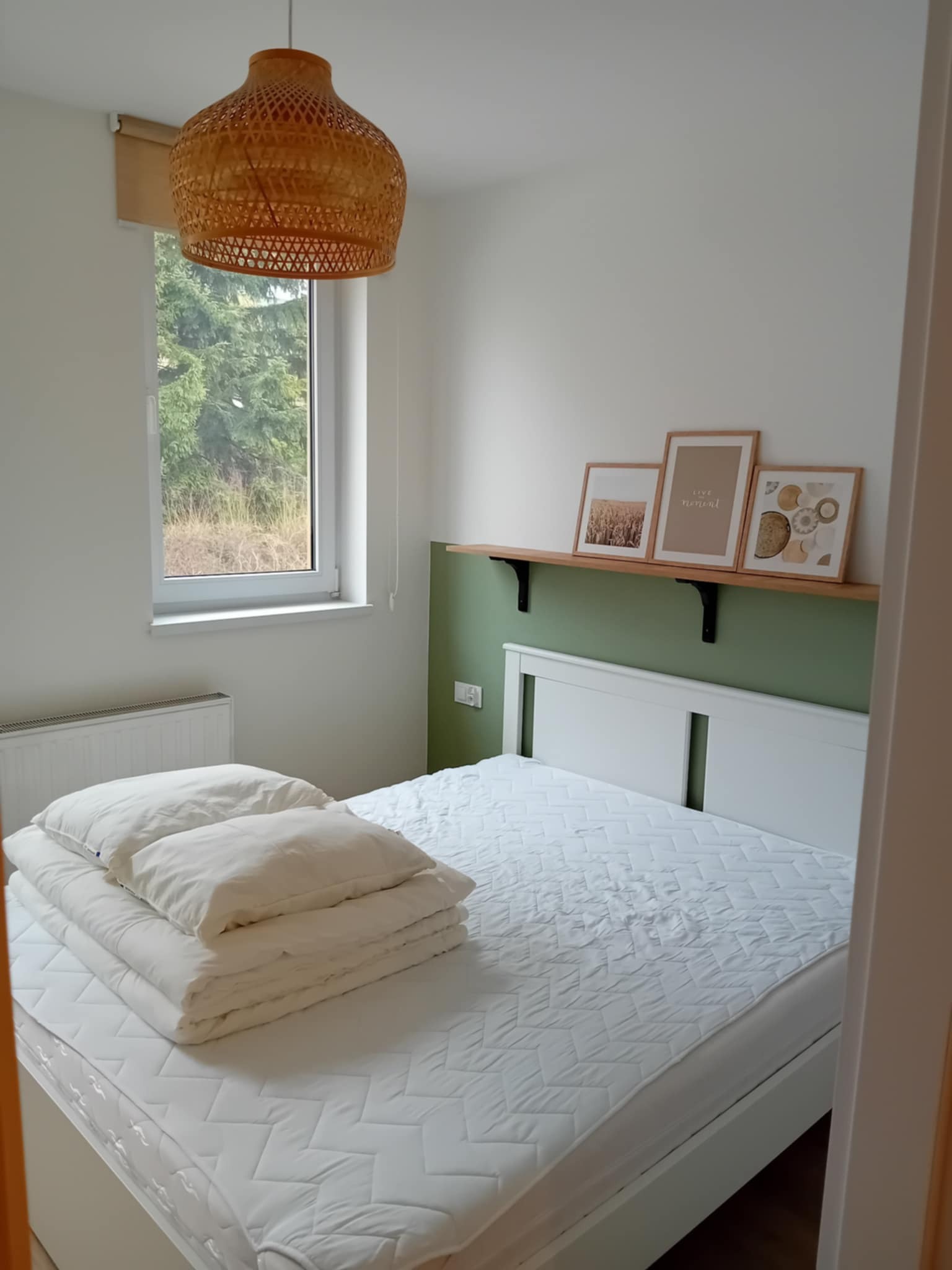 Jasna sypialnia z białym łóżkiem, drewnianą półką i zielonym akcentem na ścianie. W oknie widok na drzewa, a nad łóżkiem wisi pleciona lampa.
