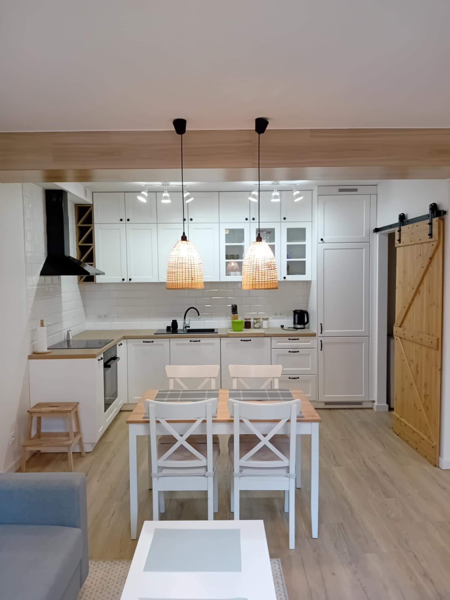 Jasna kuchnia z białymi meblami, drewnianym blatem i przesuwnymi drzwiami do spiżarni. Nad stołem wiszą dwie lampy z abażurami z wikliny.