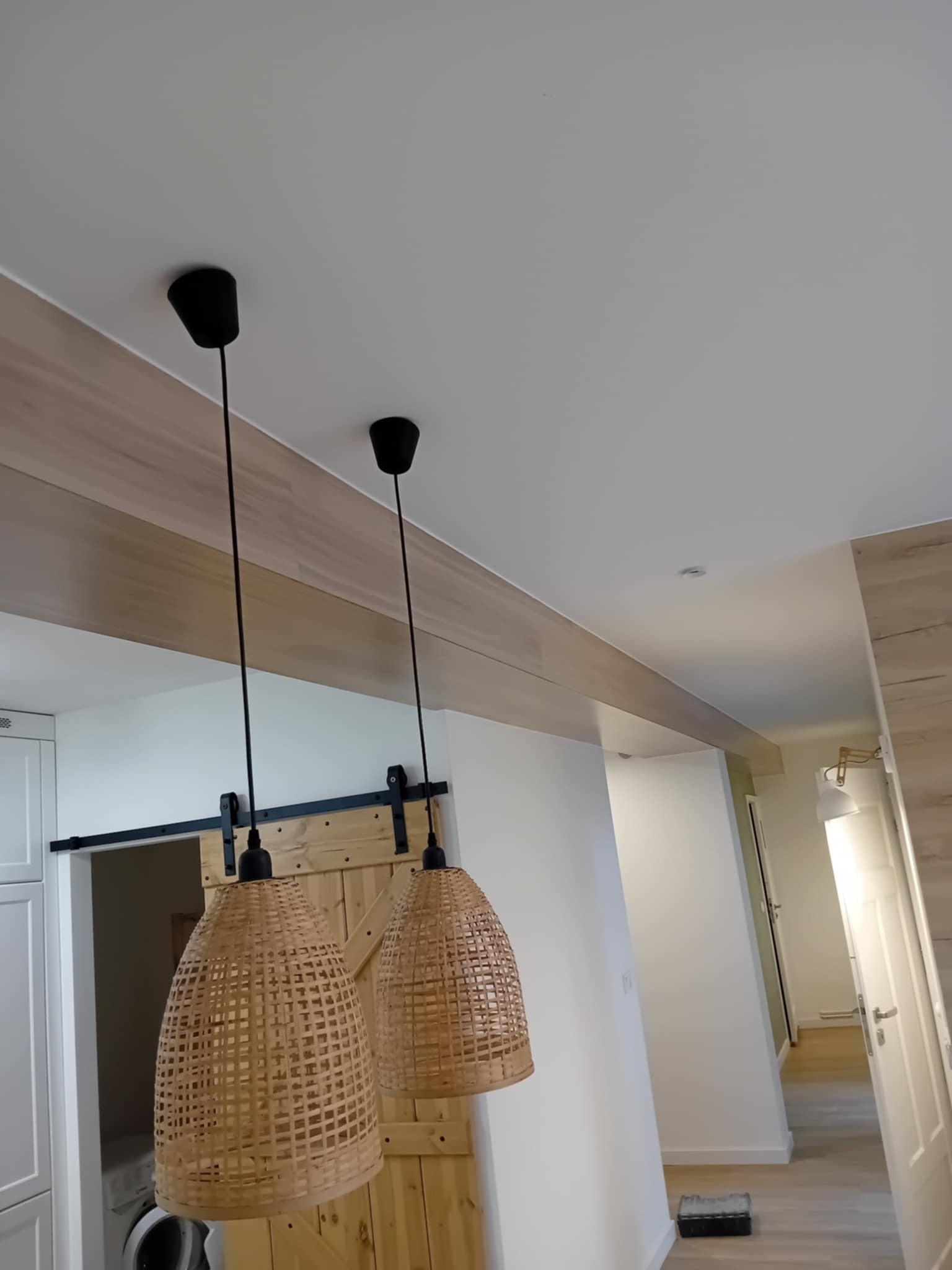 Dwa wiklinowe abażury wiszą na tle drewnianych drzwi przesuwnych i białych szafek. Widoczny fragment korytarza z jasnymi ścianami i drzwiami.