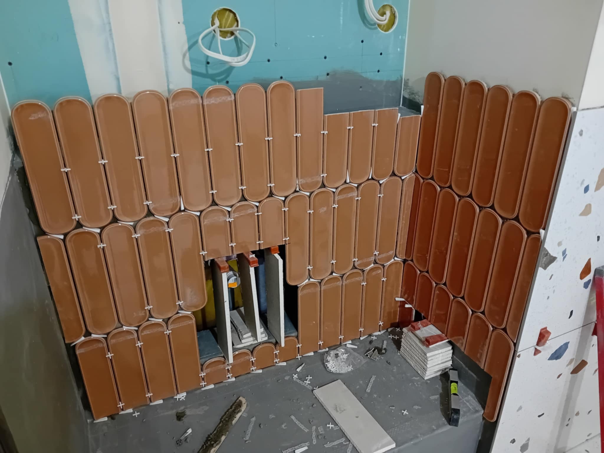 Układanie podłużnych, brązowych płytek w łazience. Widoczne krzyżyki dystansowe i fragmenty płytek. Etap prac wykończeniowych na ścianie.