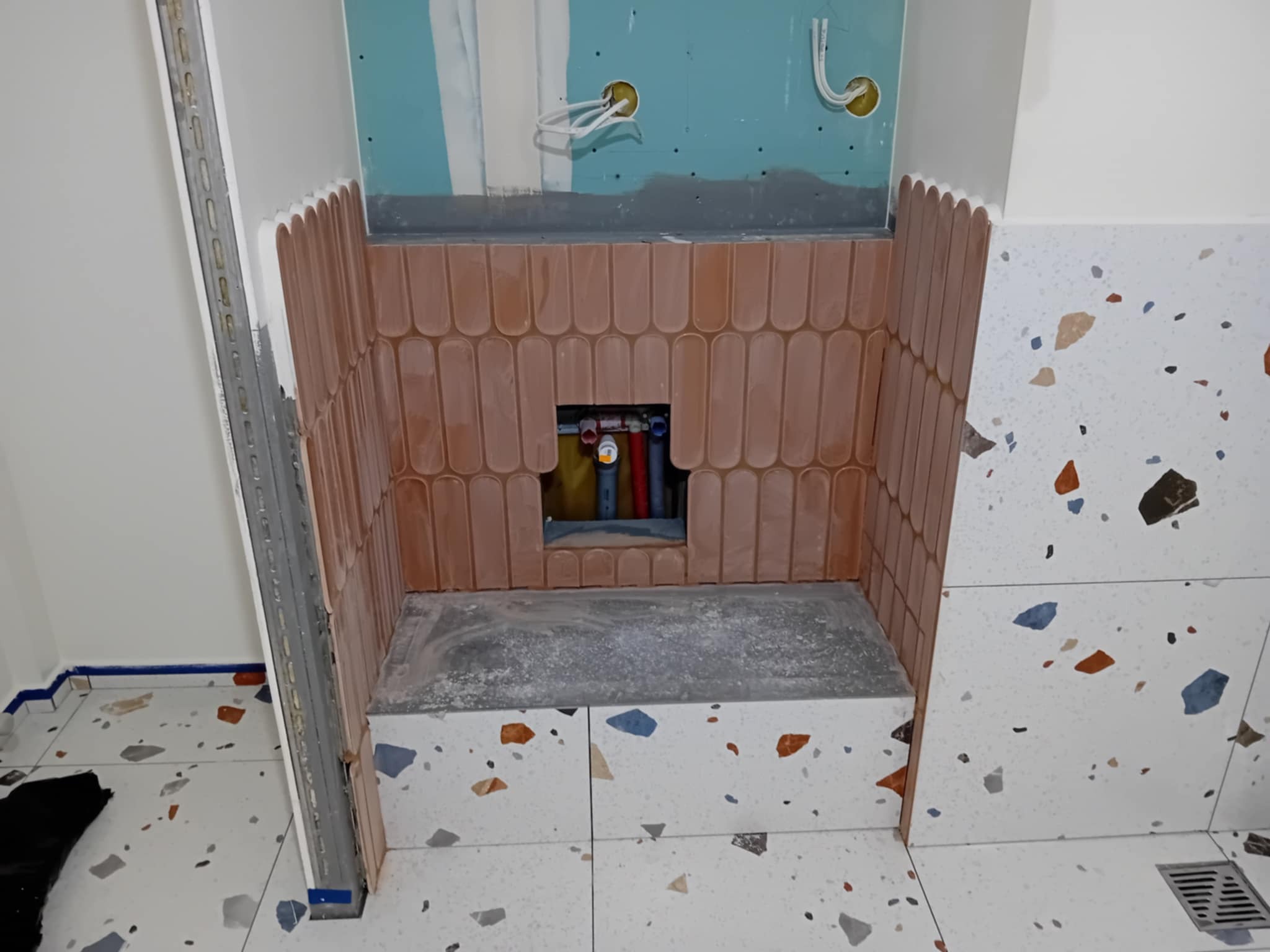 Narożnik łazienki w trakcie wykańczania: ułożone cegiełki i terazzo, widoczne rury i instalacja elektryczna. Metalowy profil w rogu.