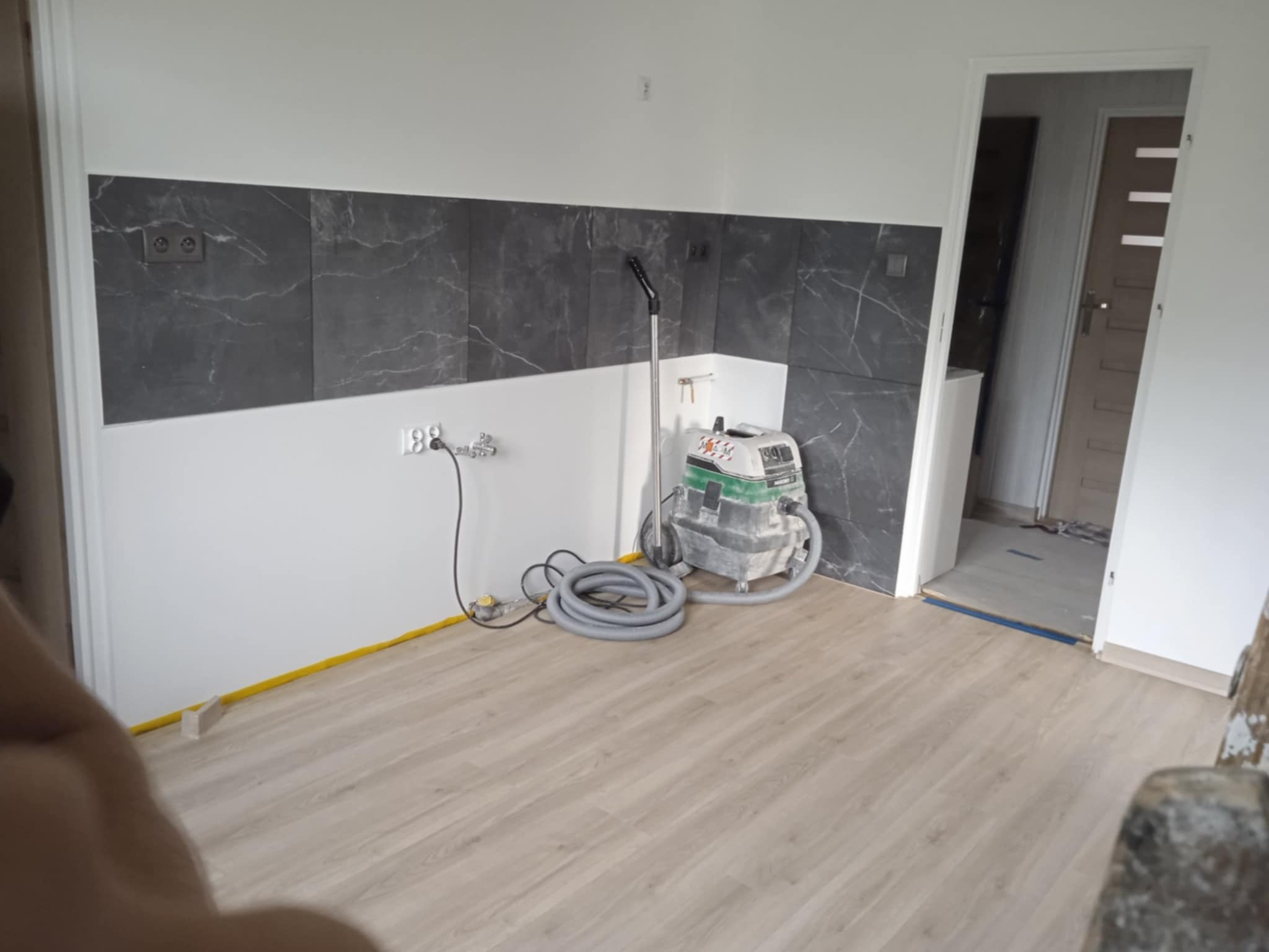 Kuchnia w trakcie prac wykończeniowych: położone płytki imitujące marmur, zabezpieczona podłoga, odkurzacz przemysłowy. Widok na otwarte drzwi do innego pomieszczenia.