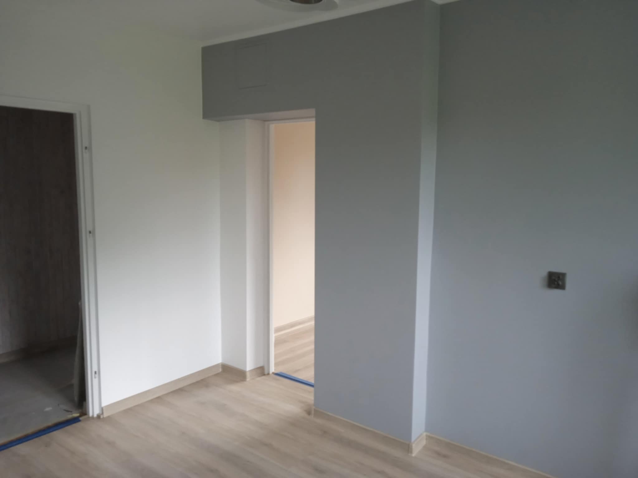 Wnętrze po malowaniu: szara ściana z kontaktem, biała ściana, drzwi w tle i panele podłogowe. Widoczny fragment sufitu z oświetleniem.