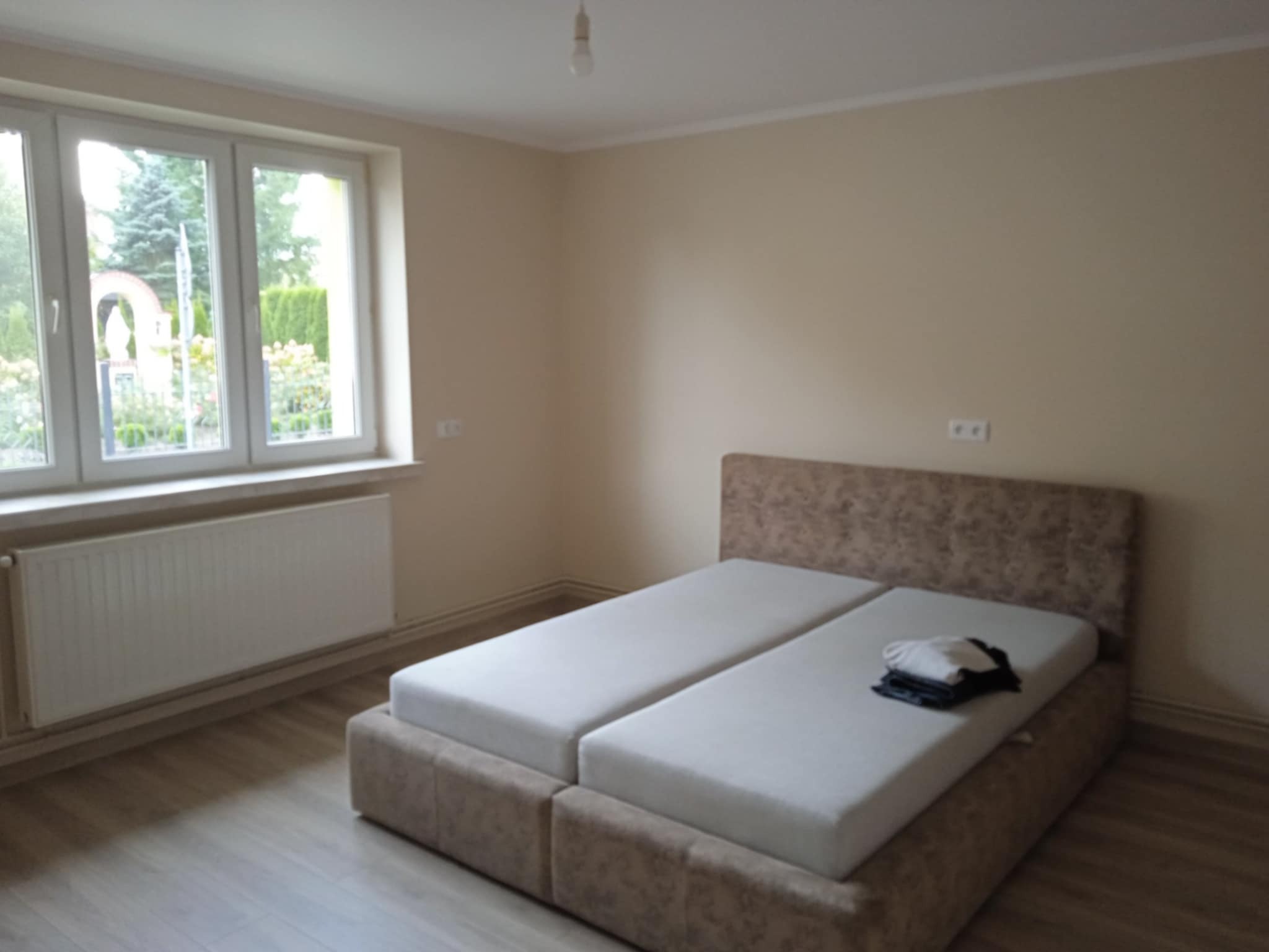 Jasne wnętrze sypialni z podwójnym łóżkiem i oknem wychodzącym na zieleń. Ściany w odcieniach beżu, drewnopodobna podłoga. Minimalistyczny styl.