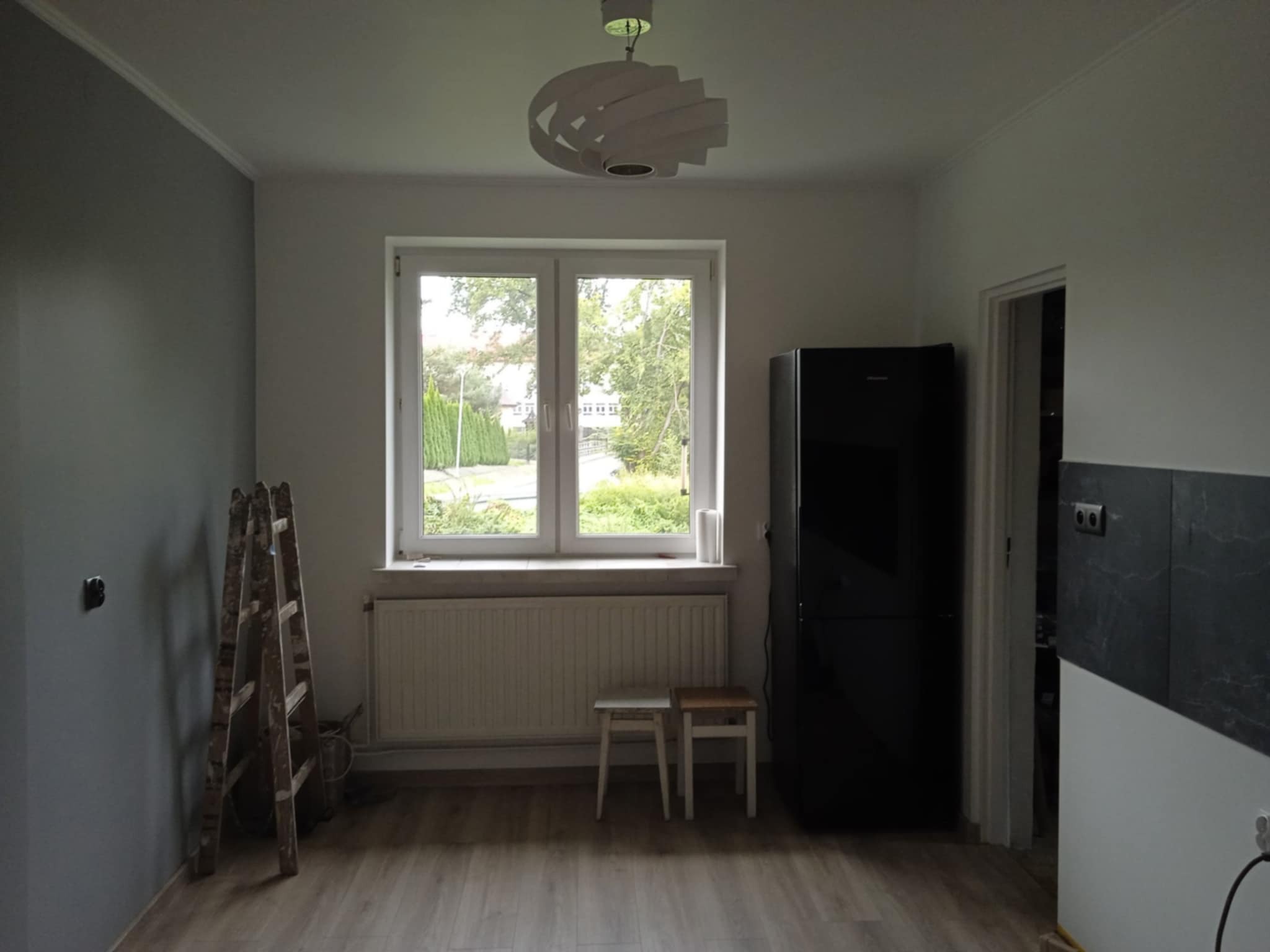 Wnętrze kuchni w trakcie remontu: drewniana drabina oparta o szarą ścianę, okno z widokiem na zieleń, czarna lodówka, krzesła. W tle widoczne płytki na ścianie.