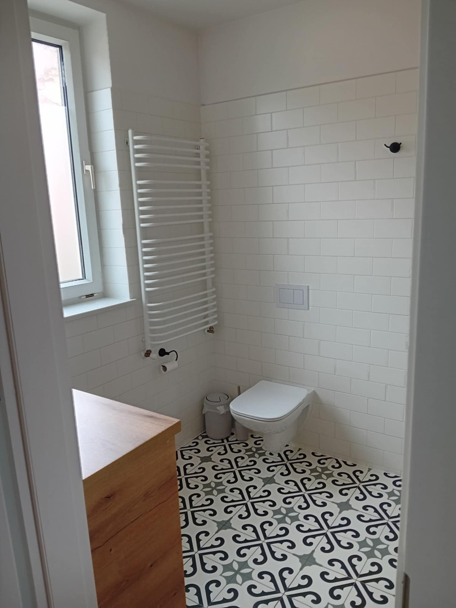 Wykończona, jasna łazienka z białą armaturą, grzejnikiem drabinkowym, wzorzystą podłogą i drewnianą szafką pod umywalkę, widok zza otwartych drzwi.