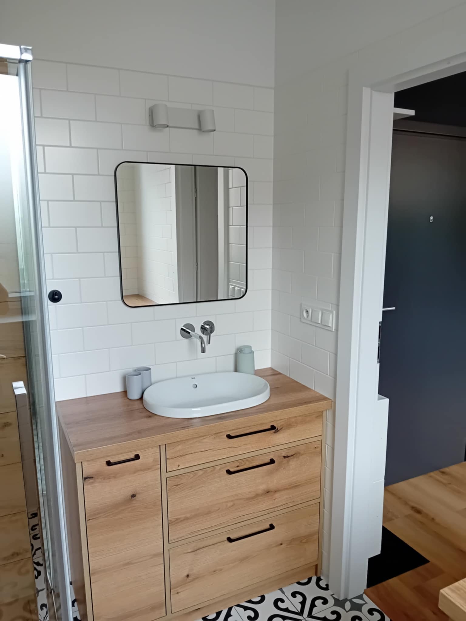 Nowoczesna łazienka z białymi płytkami, drewnianą szafką pod umywalką i czarnym lustrem. Geometryczna podłoga. Widoczny fragment kabiny prysznicowej.