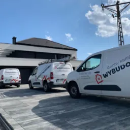 Trzy białe busy firmy dekarskiej z logo na tle nowoczesnego domu z ciemnym dachem i błękitnego nieba. Jeden z busów ma drabinę na dachu. Kostka brukowa na podjeździe.