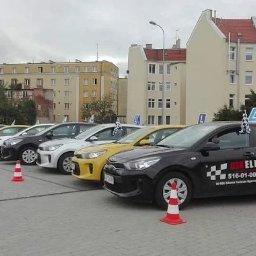 OŚRODEK SZKOLENIA KIEROWCÓW ELITE ARTUR OLSZEWSKI - Flota samochodów szkoły jazdy na parkingu miejskim, z oznaczeniami 'L' i stożkami. Dominują kolory: czarny, żółty, biały i srebrny. W tle budynki mieszkalne.