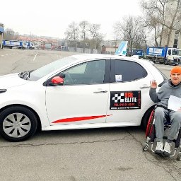 OŚRODEK SZKOLENIA KIEROWCÓW ELITE ARTUR OLSZEWSKI - Instruktor na wózku inwalidzkim pozuje obok białego samochodu z oznaczeniem 'OSK Elite', tabliczką 'L' na dachu i czerwonymi akcentami.