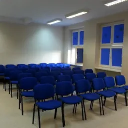 Sala szkoleniowa z rzędami niebieskich krzeseł, jasne ściany, okna z niebieskimi roletami i oświetlenie sufitowe. Gotowa do przeprowadzenia kursu.