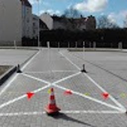 OŚRODEK SZKOLENIA KIEROWCÓW ELITE ARTUR OLSZEWSKI - Plac manewrowy szkoły jazdy z wyznaczonymi liniami i pachołkami, widok z perspektywy kursanta, słoneczny dzień, błękitne niebo, budynek w tle.