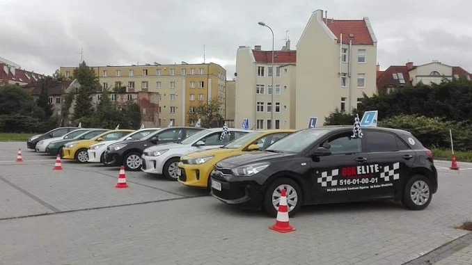 Flota samochodów szkoły jazdy na parkingu miejskim, z oznaczeniami 'L' i stożkami. Dominują kolory: czarny, żółty, biały i srebrny. W tle budynki mieszkalne.