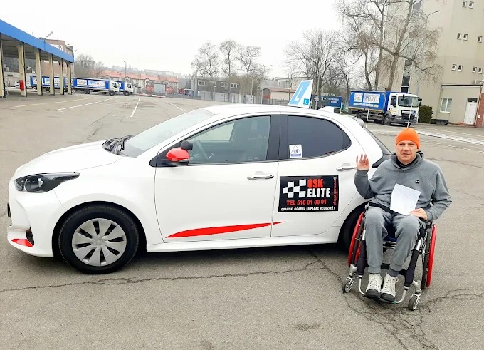 Instruktor na wózku inwalidzkim pozuje obok białego samochodu z oznaczeniem 'OSK Elite', tabliczką 'L' na dachu i czerwonymi akcentami.