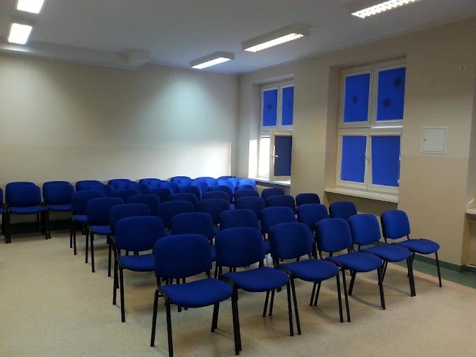 Sala szkoleniowa z rzędami niebieskich krzeseł, jasne ściany, okna z niebieskimi roletami i oświetlenie sufitowe. Gotowa do przeprowadzenia kursu.
