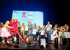 Wręczenie nagrody - roweru, podczas uroczystości z maskotką i publicznością na scenie. Osoba w garniturze przemawia, a dzieci odbierają nagrody.