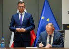 Dwóch mężczyzn w garniturach, jeden podpisuje dokument, drugi stoi z tyłu, flaga UE w tle. Butelka wody na stole. Oświetlenie studyjne z lekkim cieniem.