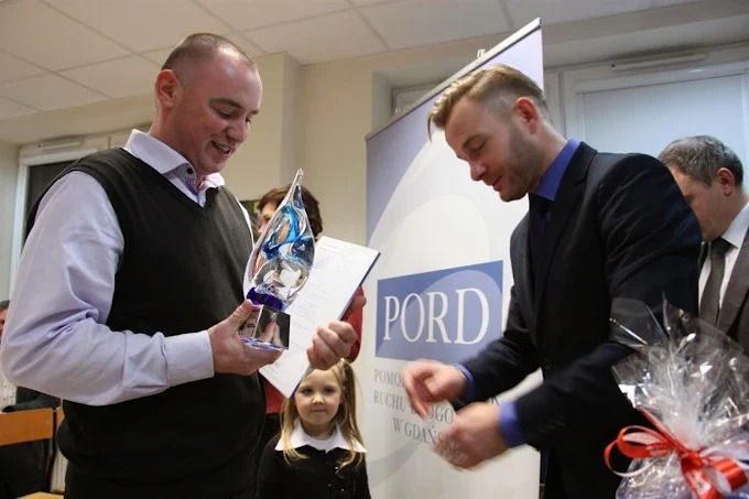 Wręczenie nagrody w kształcie płomienia przez PORD w Gdańsku. Mężczyzna w garniturze podpisuje dokument, a drugi trzyma statuetkę. W tle dziecko.