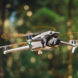 Marek Wrona - Szary dron DJI Mavic 3 w locie na tle rozmytego, zielonego lasu. Wirujące śmigła tworzą pomarańczowe aureole. Widoczna kamera drona z przodu.