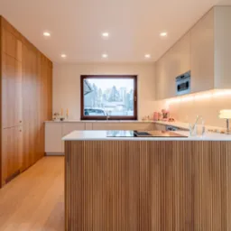 Nowoczesna kuchnia z drewnianą zabudową i wyspą z ryflowanym frontem. Jasne blaty i subtelne oświetlenie LED. Widok na zimowy krajobraz za oknem.