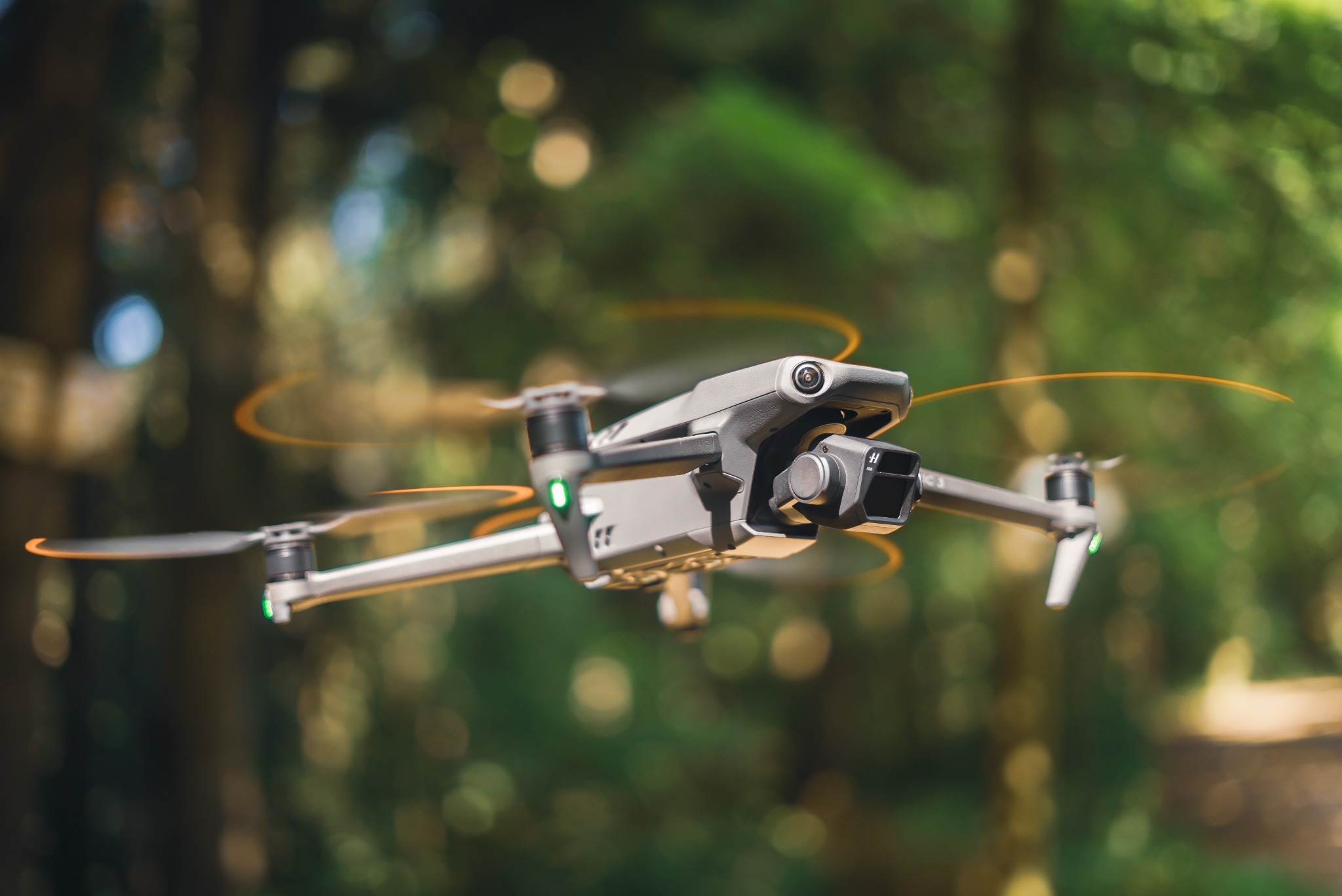 Szary dron DJI Mavic 3 w locie na tle rozmytego, zielonego lasu. Wirujące śmigła tworzą pomarańczowe aureole. Widoczna kamera drona z przodu.