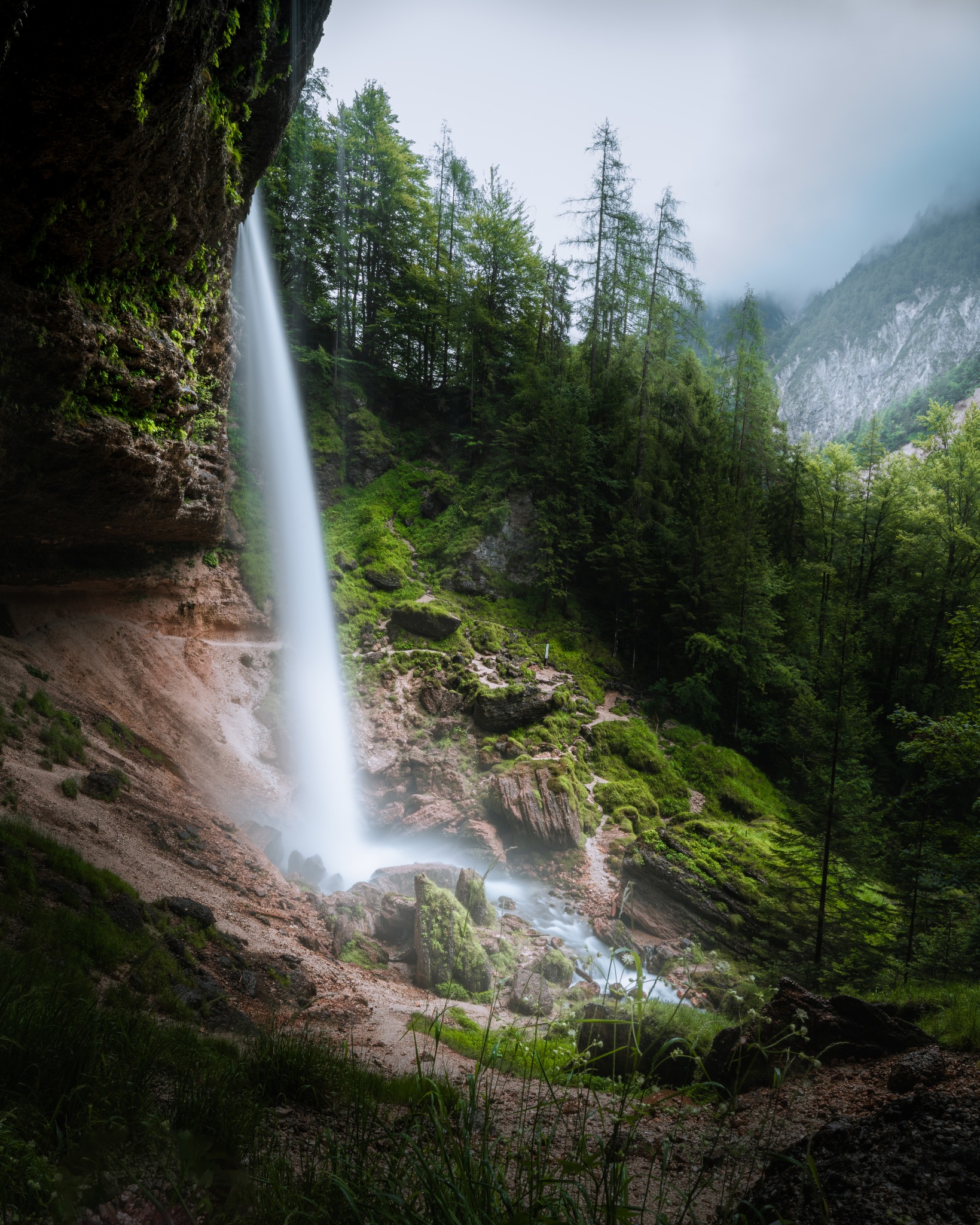 Malowniczy wodospad spływający z jaskini, otoczony zielenią lasu i mglistymi górami w tle. Ujęcie z dołu, podkreślające potęgę natury i piękno krajobrazu.
