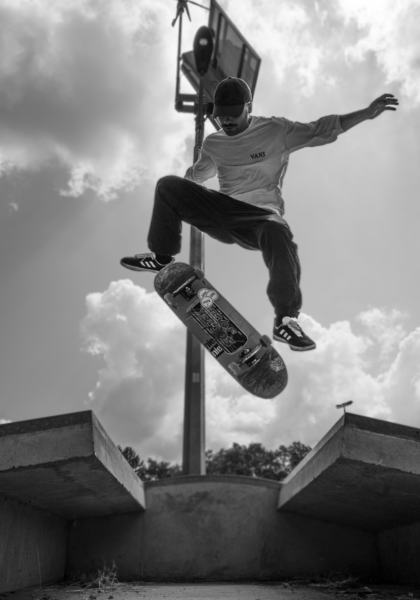 Czarno-białe ujęcie z dołu: skater w locie nad betonowym skateparkiem, w tle pochmurne niebo i wysoki słup z lampą. Widoczne logo Vans na koszulce.