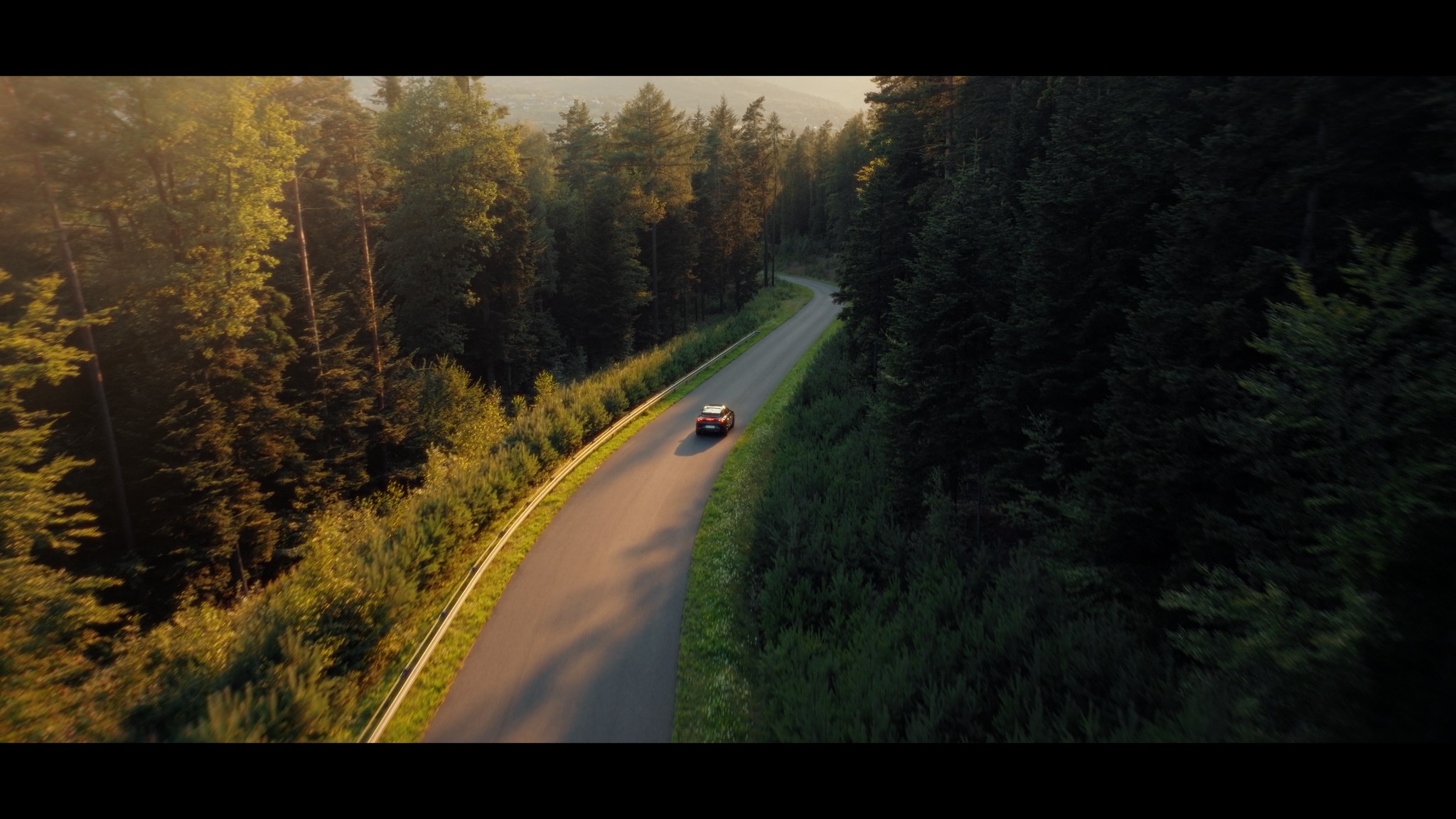 Samochód na krętej drodze otoczonej lasem, uchwycony z lotu ptaka w ciepłym, zachodzącym słońcu, tworzącym dramatyczne cienie i podkreślającym naturalne piękno krajobrazu.