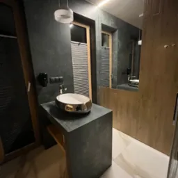 Nowoczesna łazienka z umywalką nablatową w kolorze złota, szare ściany z fakturą i oświetlenie LED. Minimalistyczny design i eleganckie wykończenie.