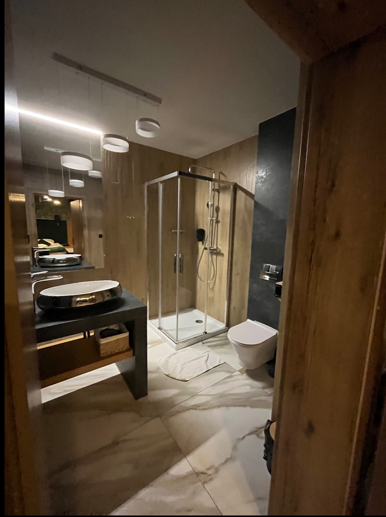 Nowoczesna łazienka z kabiną prysznicową, umywalką na blacie i marmurową podłogą. Oświetlenie punktowe podkreśla minimalistyczny design i drewniane akcenty.