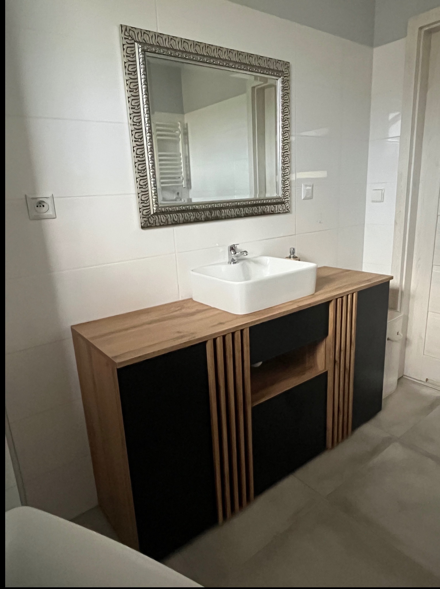 Wykończona łazienka w Grudziądzu: drewniana szafka z czarnymi frontami pod umywalką nablatową, lustro w ozdobnej ramie, białe płytki na ścianach.