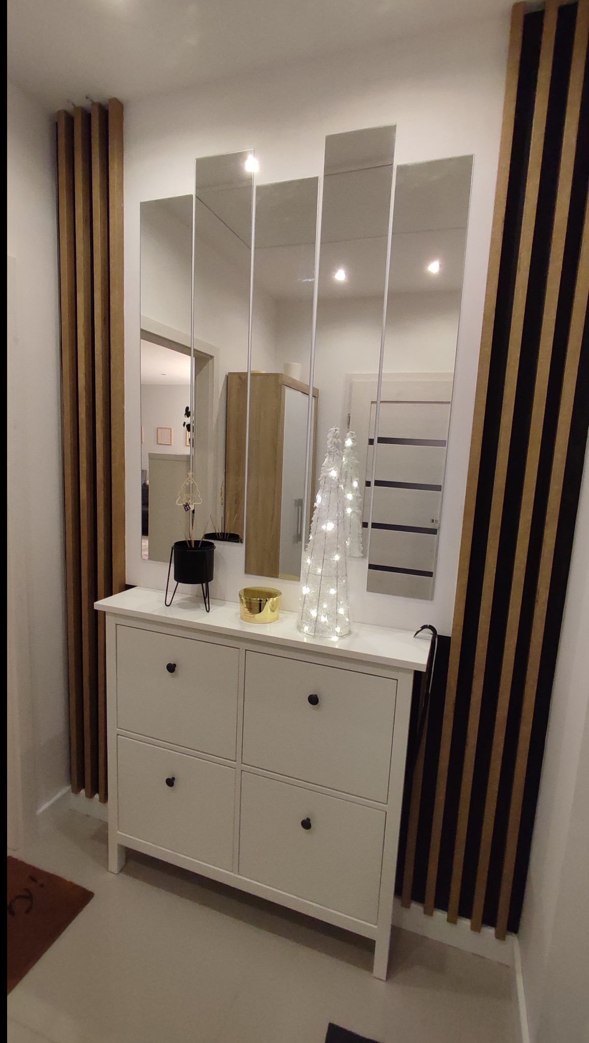 Nowoczesny przedpokój: biała szafka na buty, lustra w wąskich panelach i dekoracyjne lamele ścienne w odcieniach brązu i czerni. Minimalistyczny design.