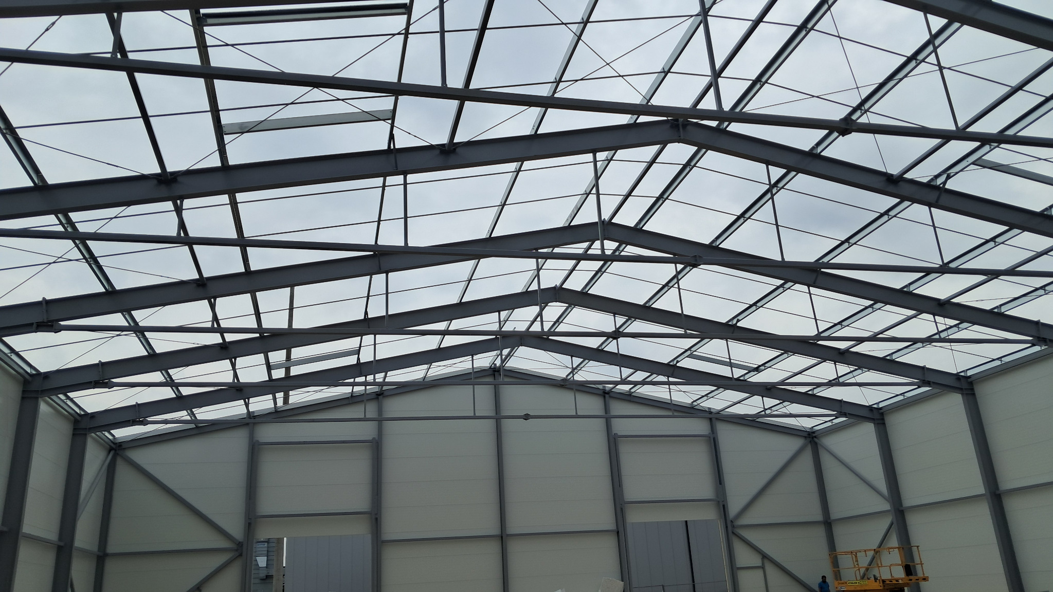Wnętrze hali w trakcie budowy; widoczne stalowe elementy konstrukcyjne dachu i ścian, jasne panele ścienne, podnośnik koszowy w rogu. Ujęcie od dołu.