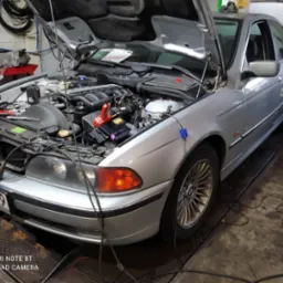 Srebrny samochód osobowy z otwartą maską w warsztacie; widoczny silnik, akumulator z klemami, narzędzia oraz przewody diagnostyczne. Auto na podnośniku.