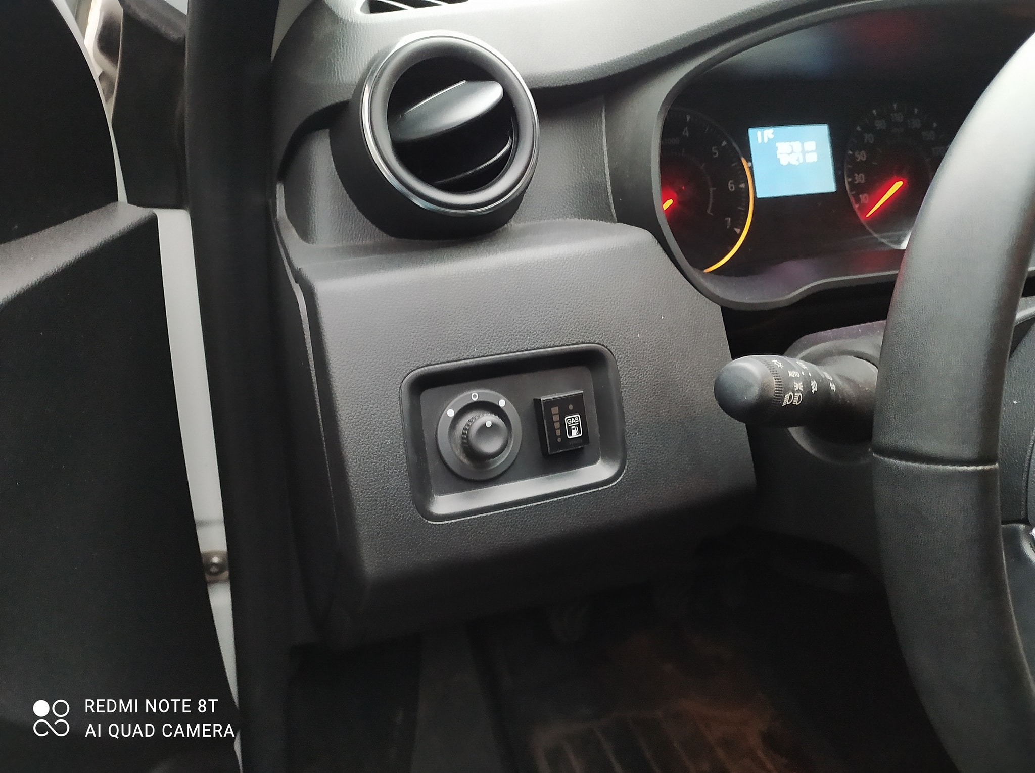 Wnętrze samochodu z zamontowanym przełącznikiem benzyna/LPG, widoczny panel sterowania i wskaźnik poziomu gazu. Element deski rozdzielczej, kierownica w kadrze.