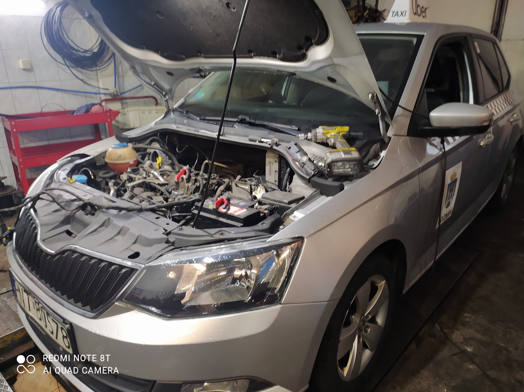 Srebrna Skoda z otwartą maską w warsztacie, widoczny silnik i narzędzia. Auto z oznaczeniami Taxi Uber. Przegląd lub naprawa w toku. Oświetlenie warsztatowe.