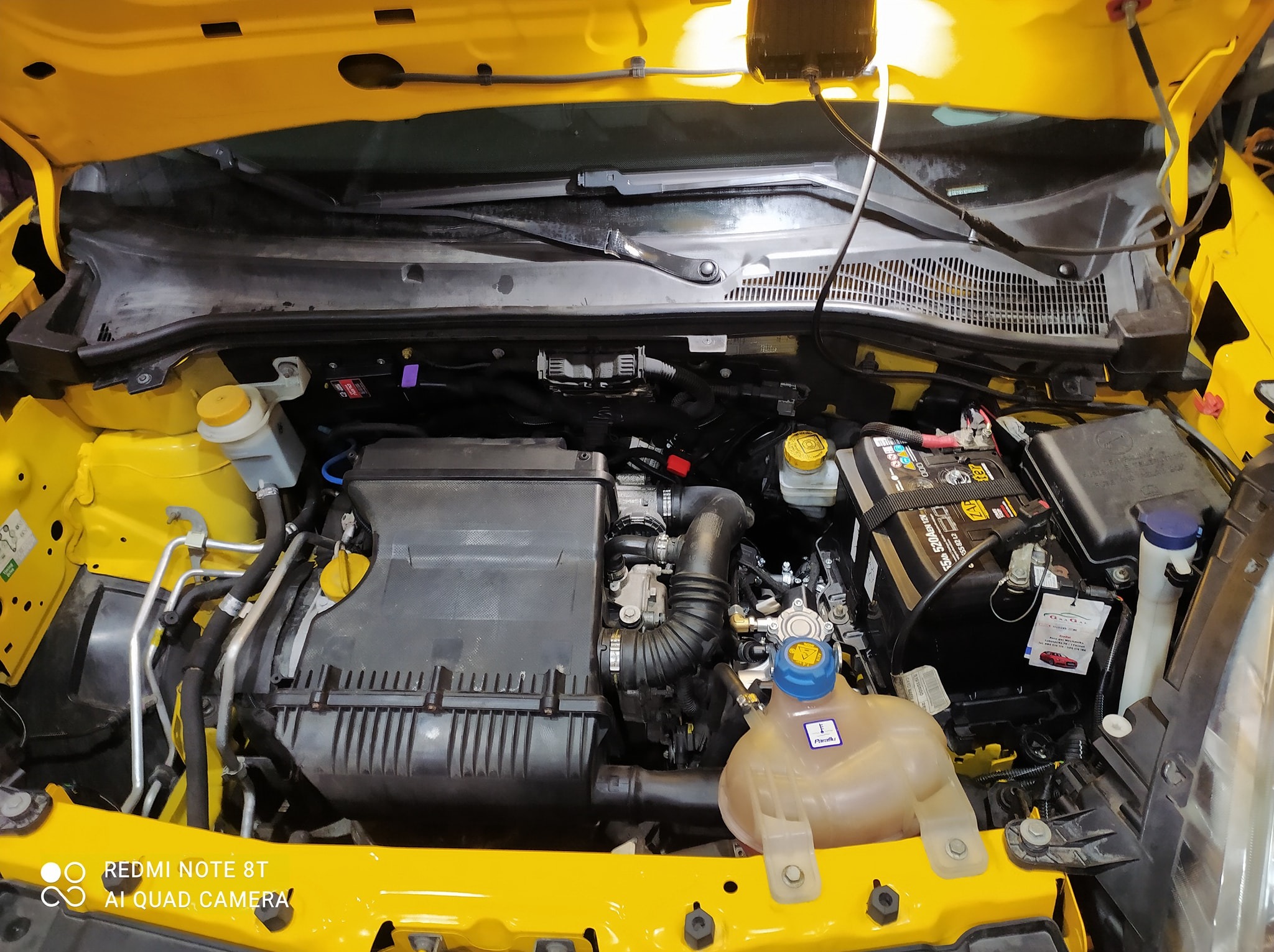 Otwarta maska żółtego samochodu, widoczny silnik z elementami instalacji LPG. Akumulator, zbiorniki płynów i przewody w komorze silnika. Oświetlenie warsztatowe.