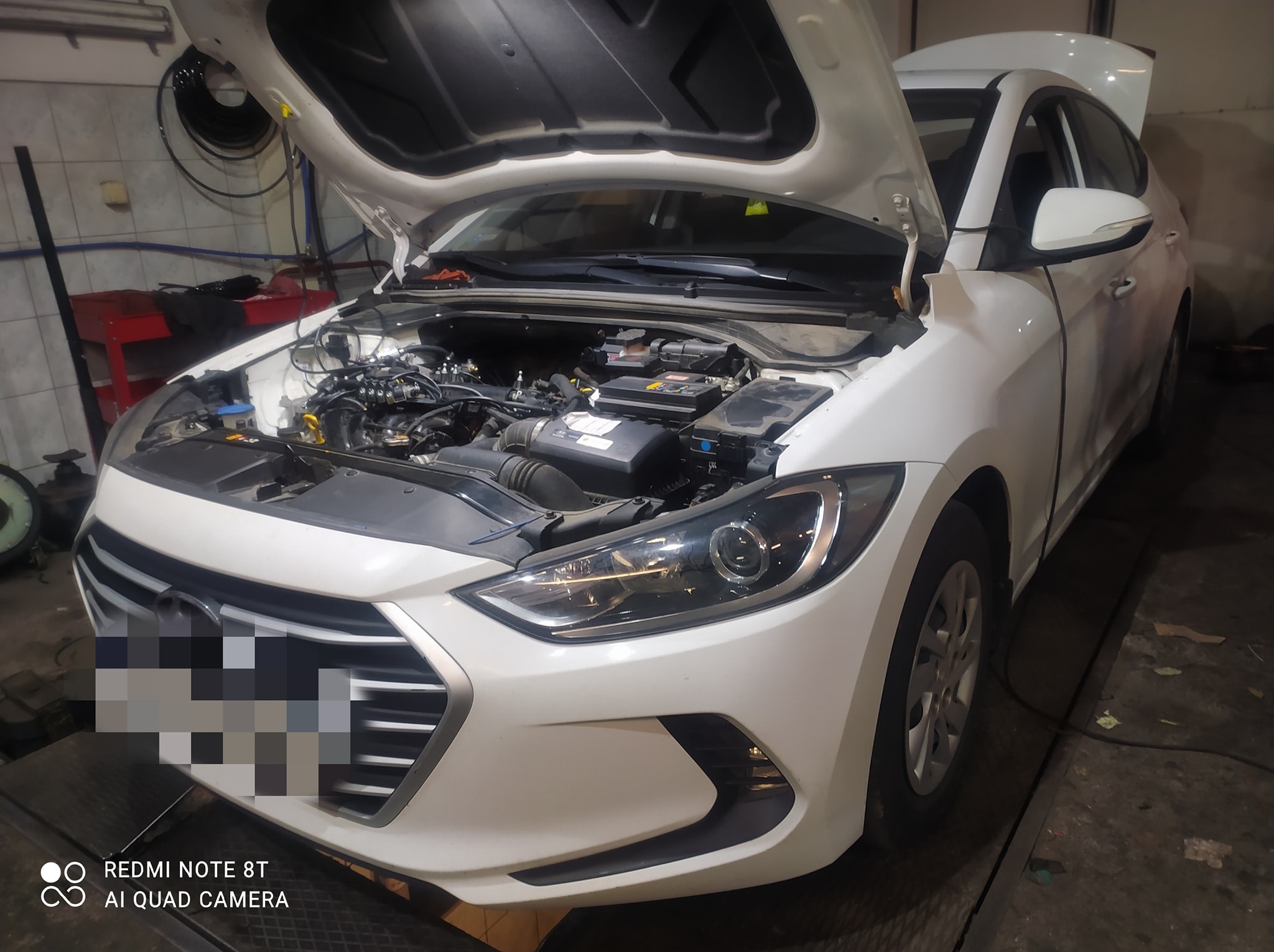 Biały samochód z otwartą maską w warsztacie. Widoczny silnik, akumulator. W tle narzędzia i wyposażenie warsztatowe.