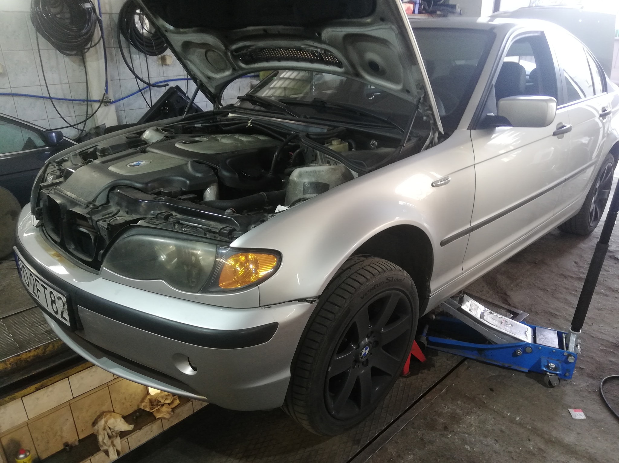 Srebrny sedan BMW z otwartą maską silnika, podnośnik hydrauliczny pod progiem, widoczny silnik i elementy osprzętu w warsztacie.