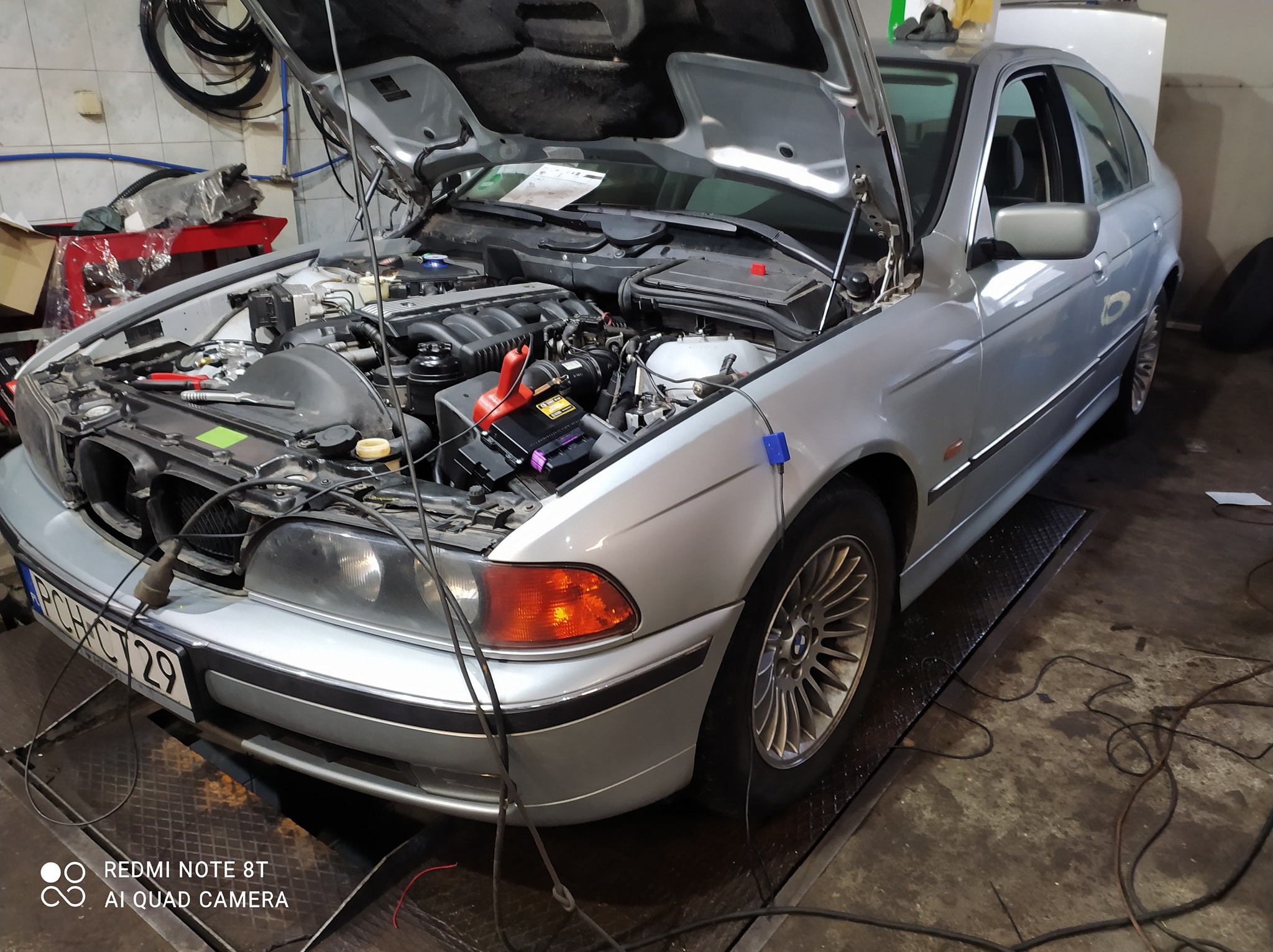 Srebrny samochód osobowy z otwartą maską w warsztacie; widoczny silnik, akumulator z klemami, narzędzia oraz przewody diagnostyczne. Auto na podnośniku.